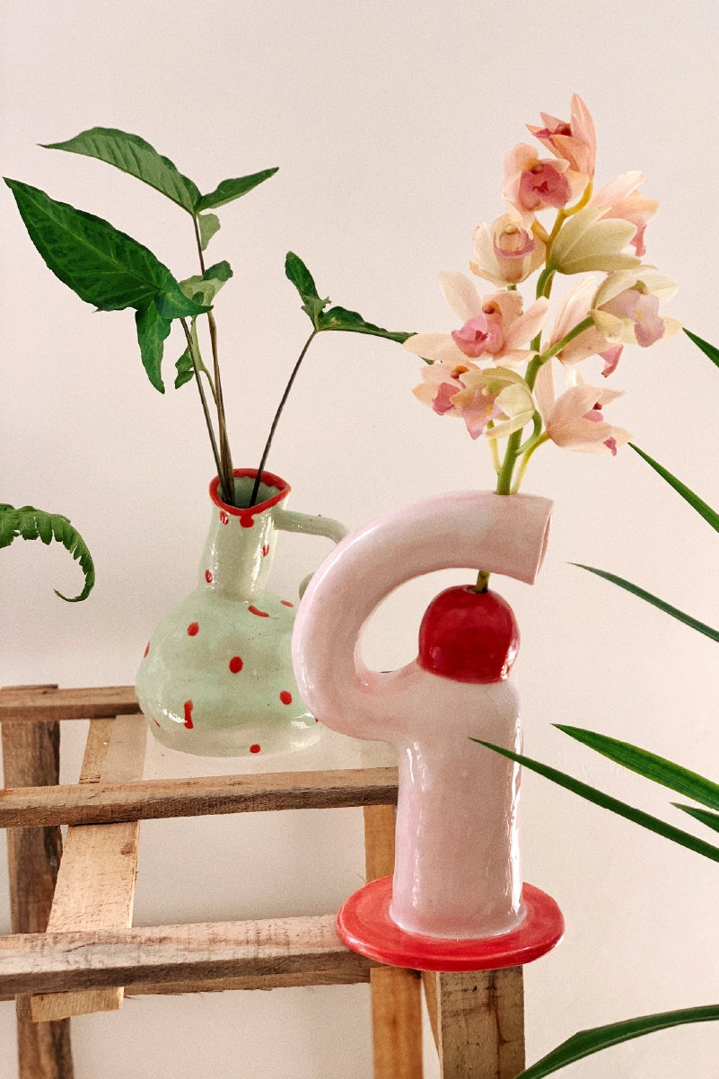 Decorative vase shaped like a letter 'F' with pink flowers on a wooden stool against a white wall.