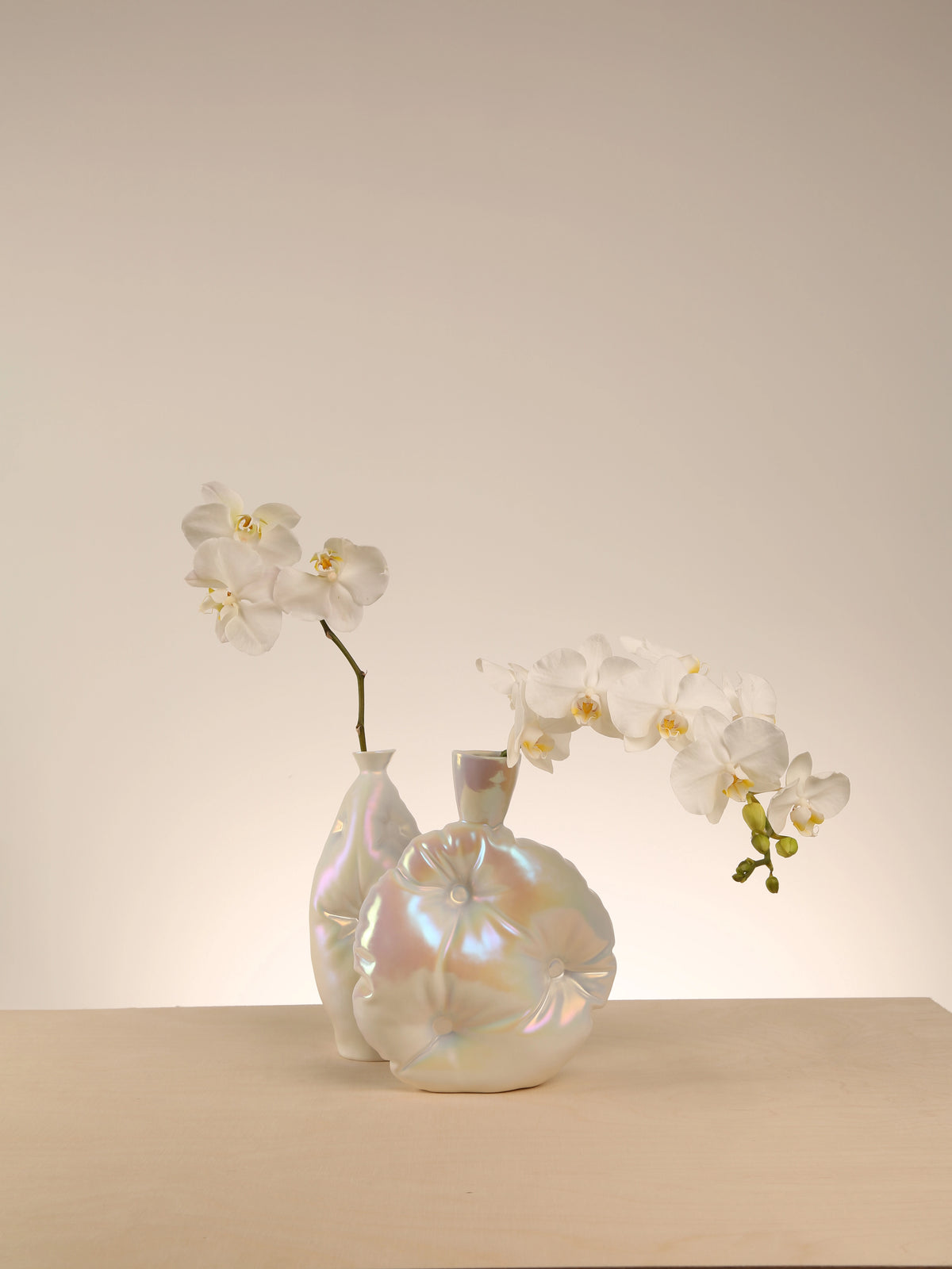 Two iridescent vases with white orchids on a beige surface and background